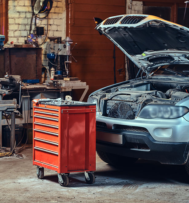 A silver car with its hood open sits beside a red five-drawer tool cart in a cluttered garage workshop.
