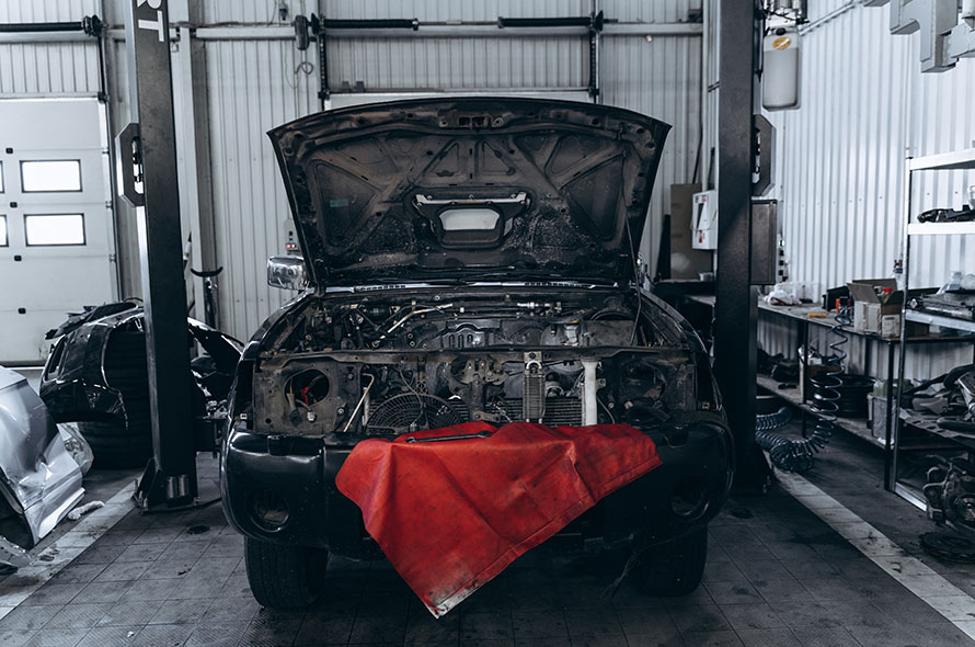 A car with its hood open in a garage, revealing the engine. A red cloth is draped over the front, surrounded by various tools and equipment.