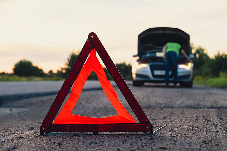A reflective warning triangle placed on the roadside, with a person inspecting the engine of a car under a sunset sky.