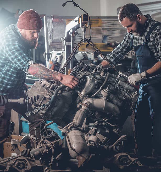 Two mechanics work together in a garage, carefully inspecting and repairing a large engine under bright workshop lights.